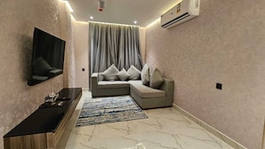 A modern living room with a sectional sofa, a flat-screen TV mounted on the wall, and an air conditioning unit. - Royal Plaza Hotel - Duqm  (Duqm)