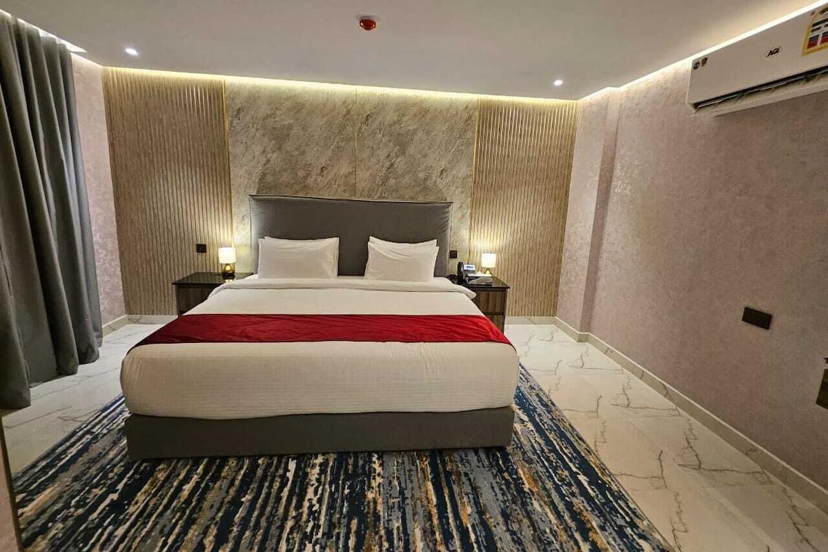 Habitación de hotel con una cama grande, dos mesitas de noche, una colcha roja, un papel tapiz con diseño de mármol y un equipo de aire acondicionado.