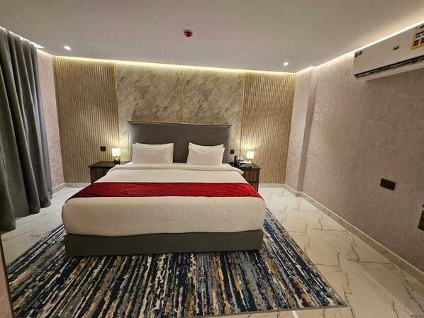 Habitación de hotel con una cama grande, dos mesitas de noche, una colcha roja, un papel tapiz con diseño de mármol y un equipo de aire acondicionado.