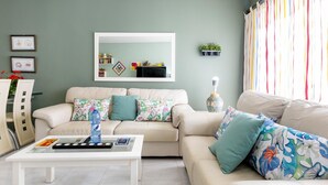Una sala de estar con un sofá beige, una mesa de centro y un televisor. Hay un espejo en la pared, una planta y una ventana con cortinas.
