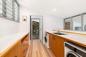A compact kitchen with a washing machine, wooden cabinets, and a window with blinds.