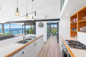 A modern kitchen with a large island, a gas stove, and a view of the ocean. - Award-Winning Oceanfront Glass Home | Sleeps 8 (Forresters Beach)