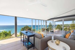 A modern living room with a large glass wall overlooking the ocean, a ceiling fan, and a seating area with a coffee table. - Sandcastle by Coast Hosting (Forresters Beach)