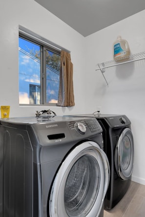 Une buanderie équipée de deux machines à laver et de deux sèche-linge à chargement frontal, d’une fenêtre donnant sur l’extérieur et d’une étagère sur laquelle se trouve une bouteille de lessive.