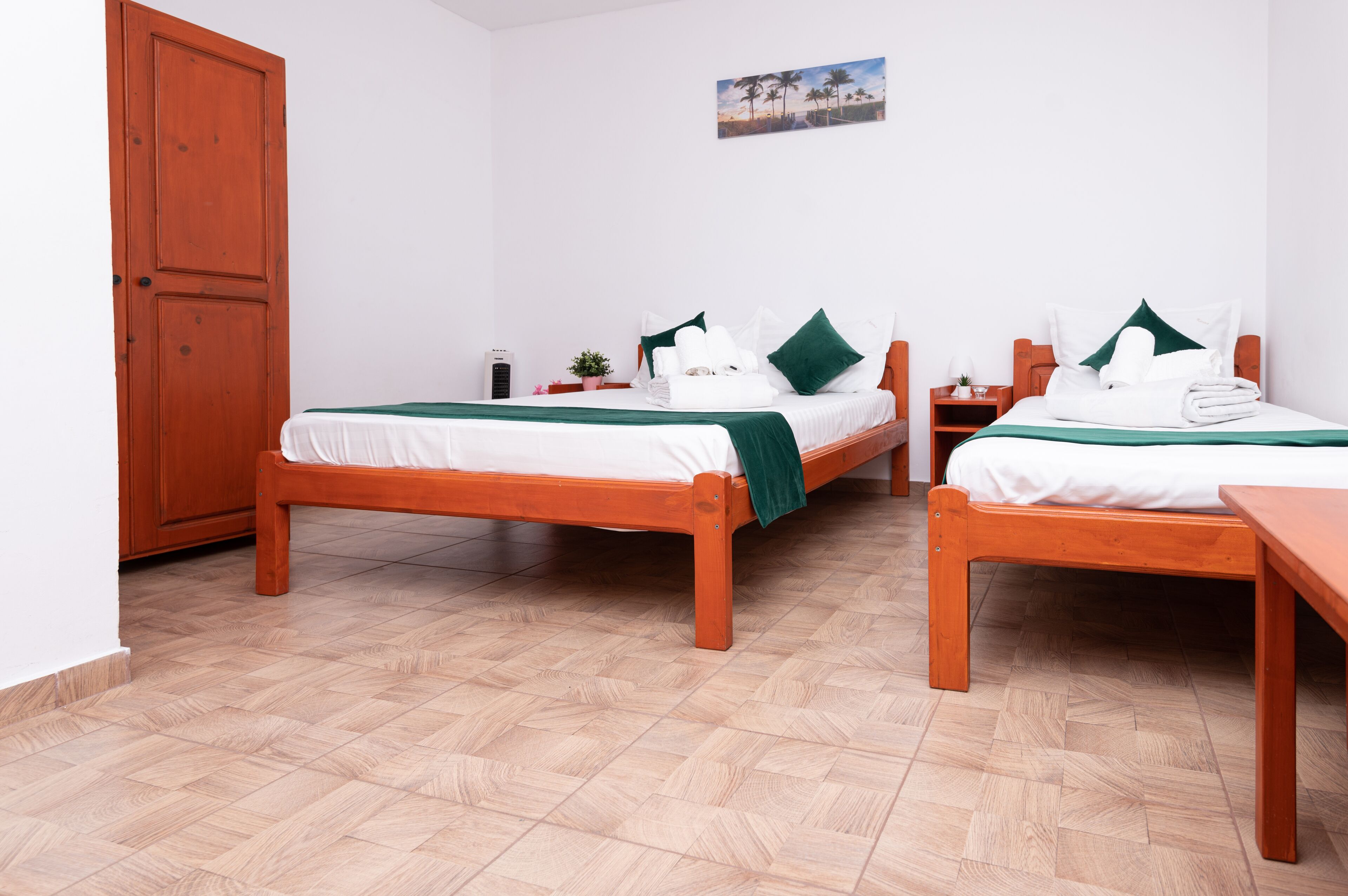 A hotel room with two wooden beds, white bedding, green pillows, a wooden floor, a wooden door, and a small table with a plant.