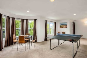 A room with a ping pong table, a table with chairs, and large windows with curtains.