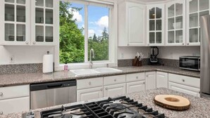 Une cuisine moderne avec des meubles blancs, un plan de travail en granit, une cuisinière à gaz, un four à micro-ondes et une vue sur la verdure à l’extérieur.