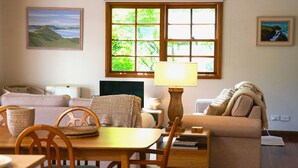 Un salon chaleureux avec une table en bois, un canapé et un téléviseur. Il y a deux fenêtres qui donnent sur un paysage verdoyant.