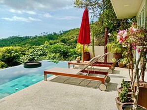 An infinity pool with lounge chairs and a red umbrella, surrounded by lush greenery and a view of a forested hill.