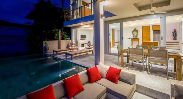 Une maison moderne dotée d’une piscine, d’un coin repas et d’un salon avec un canapé et des coussins rouges.