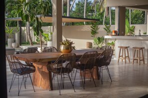 Un amplio espacio al aire libre con comedor, que incluye una gran mesa de madera rodeada de sillas de mimbre, una zona de bar con taburetes y diversas plantas en macetas.