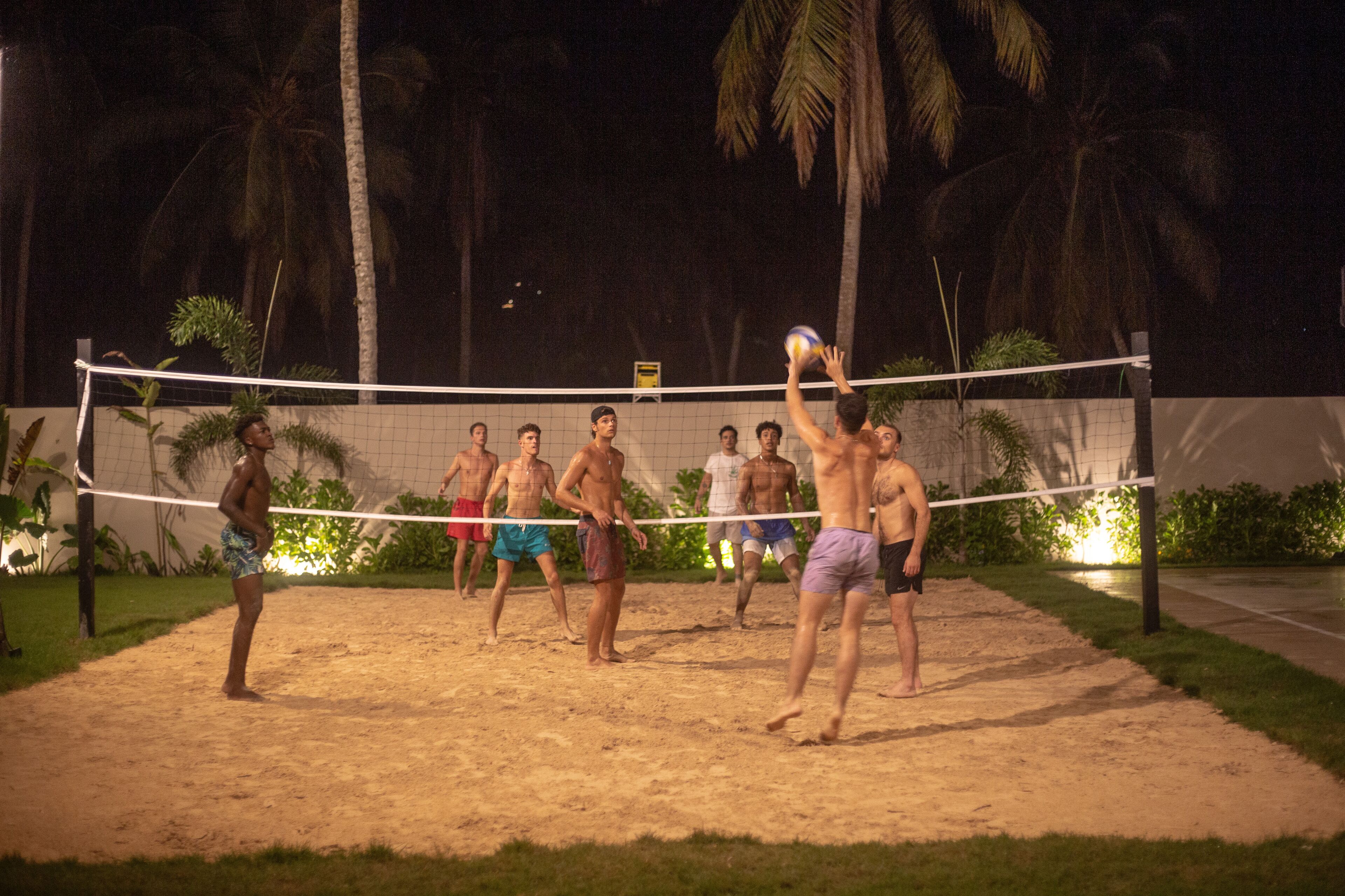 Partido nocturno de voleibol de playa con jugadores en pantalones cortos, una red de voleibol y palmeras al fondo.