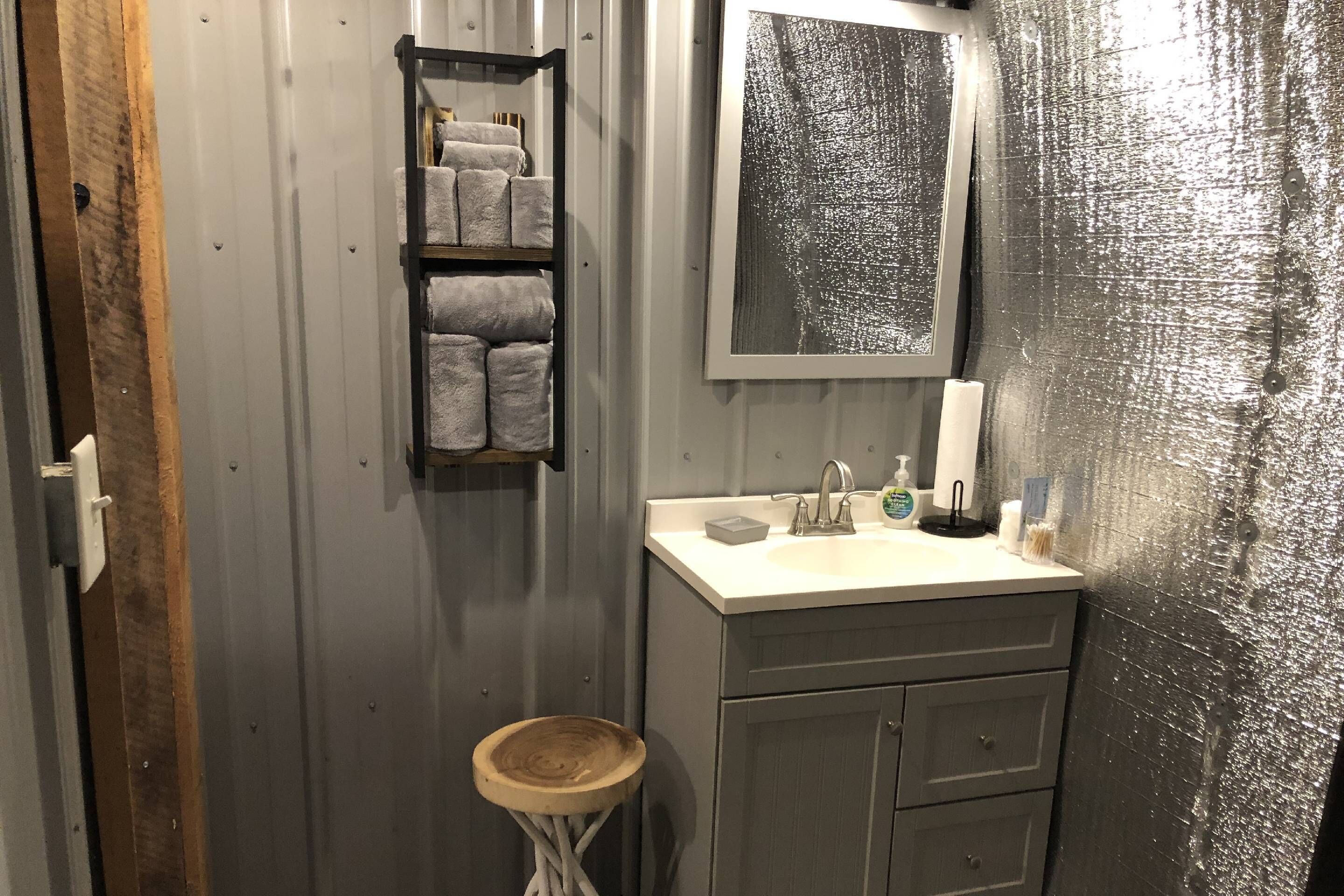 A bathroom with a wooden stool, a shelf with towels, a sink with toiletries, and a mirror.