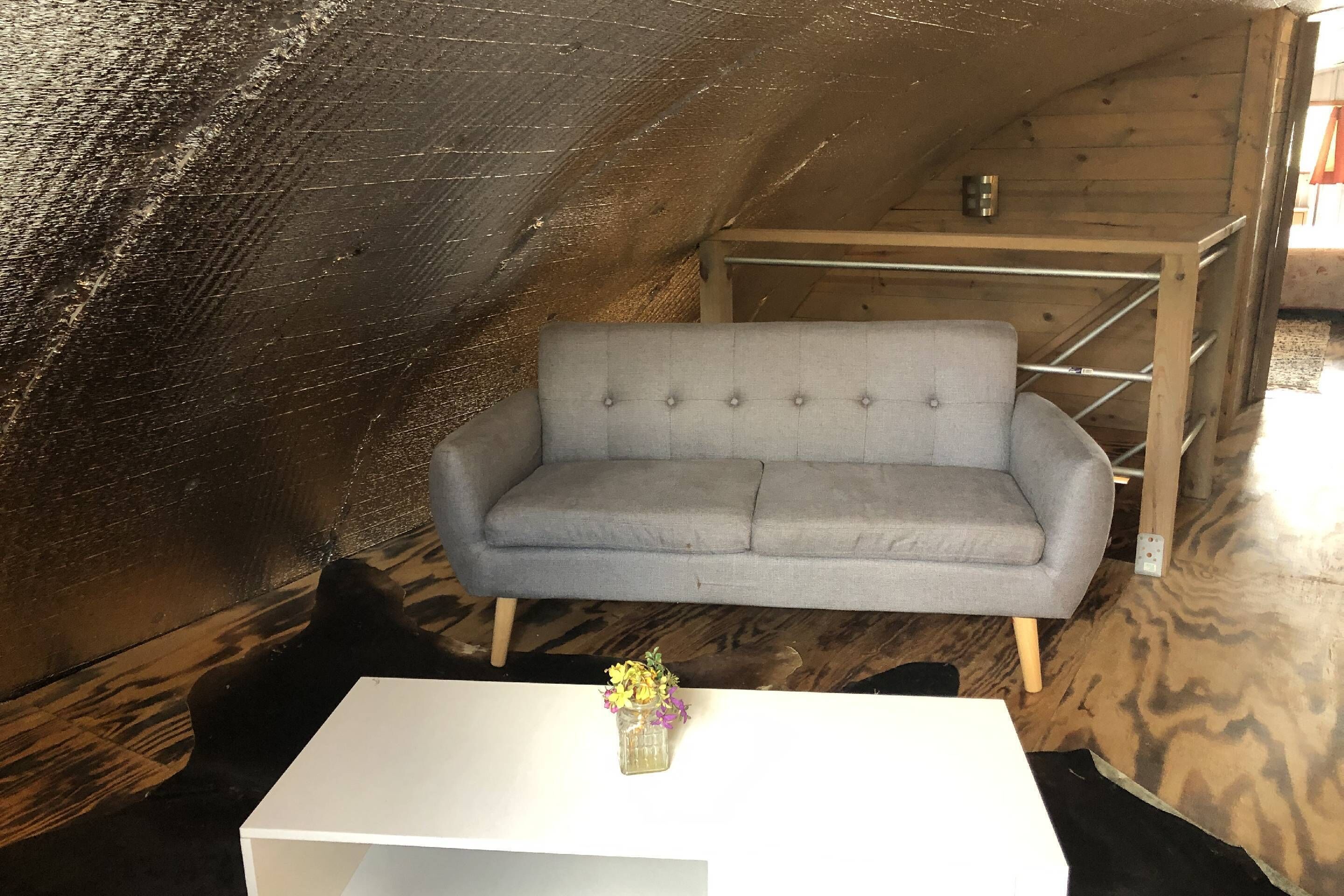 En grå sofa i et trægulvsrum med et hvidt bord og en lille vase med blomster.