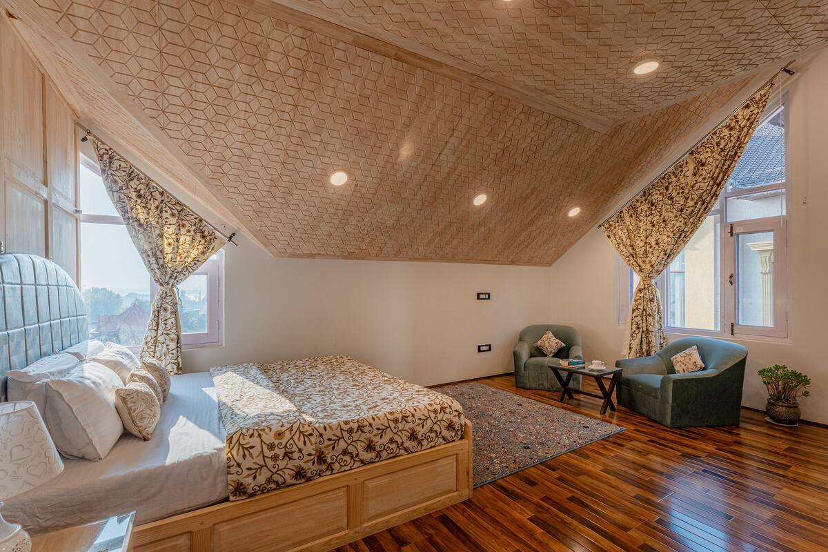 A bedroom with a sloped ceiling, a bed with patterned bedding, two chairs, a small table, and a potted plant.