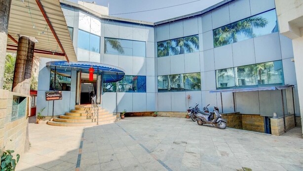 A modern building with a curved facade, large windows reflecting palm trees, and a covered entrance with steps leading up. - Saiba Residency (Paud)