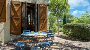 A patio with a round table and blue chairs, wooden shutters, and a view of greenery.