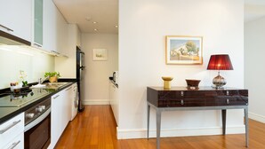 Una cocina moderna con armarios blancos, encimera negra y suelo de madera. Hay una mesa con una lámpara y un cuadro en la pared.