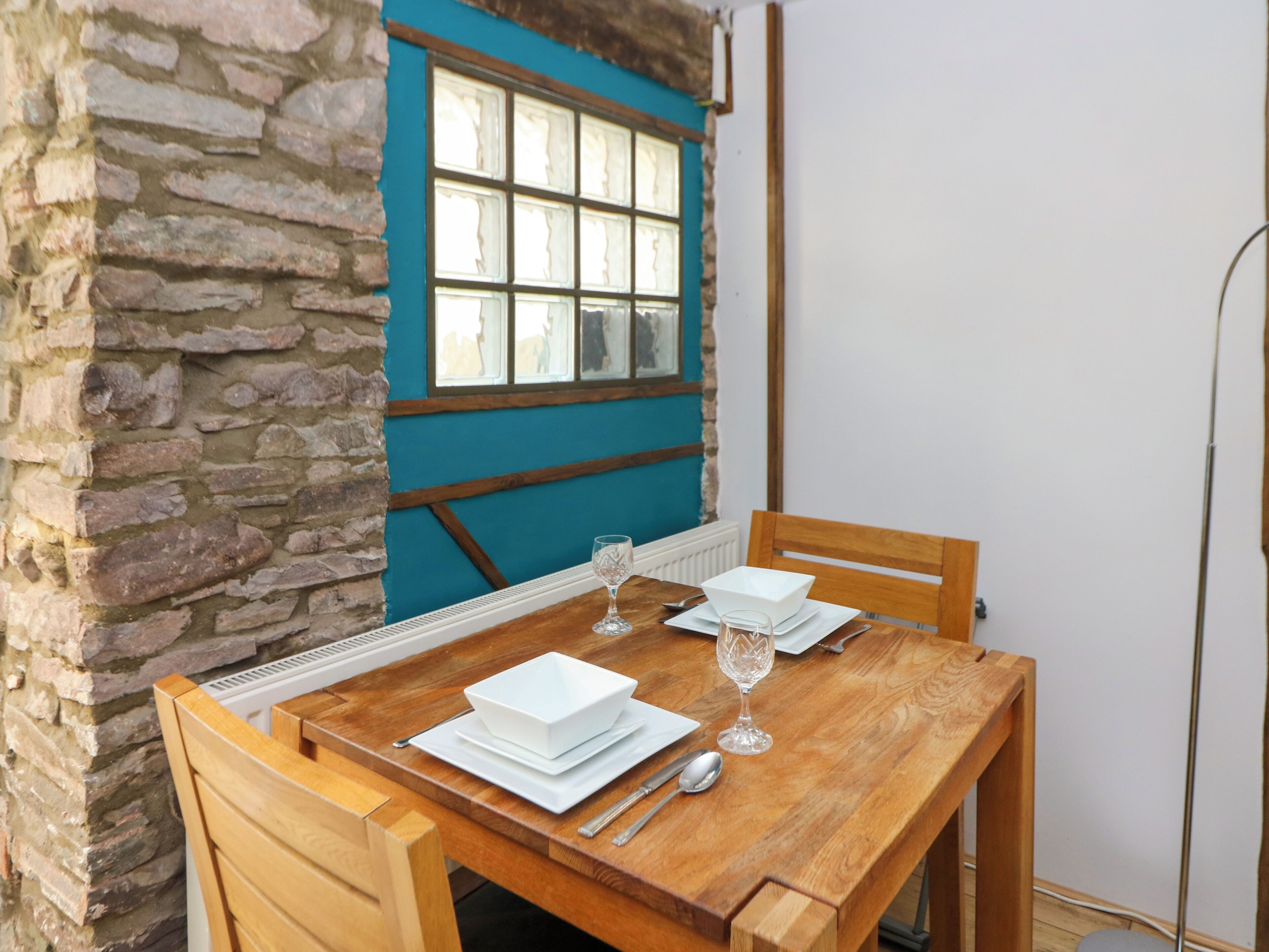 A wooden dining table with white plates, a wine glass, and cutlery. The table is set against a stone wall and a window with blue trim.
