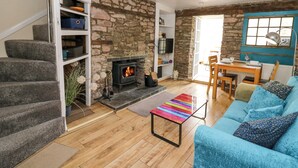 A cozy living room with a stone wall, a fireplace, a staircase, a dining area, and a blue sofa. - Rhif Deg (Brecon)