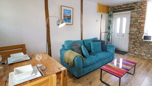A cozy living room with a wooden dining table set for two, a teal sofa with cushions, a colorful striped coffee table, and a stone wall. - Rhif Deg (Brecon)