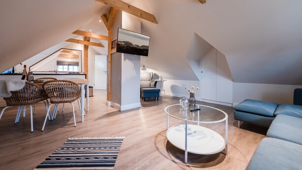 A spacious loft apartment with high ceilings, exposed wooden beams, a dining area with wicker chairs, a round coffee table, and a blue sofa. - Appartement Wattside (Morsum)
