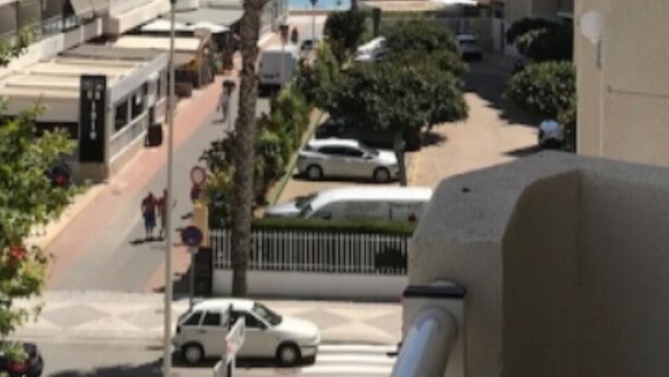Une vue depuis un balcon donnant sur une rue bordée de palmiers, avec un passage piéton et un immeuble doté d’un balcon.