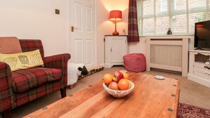 Ein gemütliches Wohnzimmer mit einem karierten Sofa, einem Holztisch mit Obstschale, einem weißen Schrank, einer roten Lampe und einem Fernseher auf einem Lowboard.