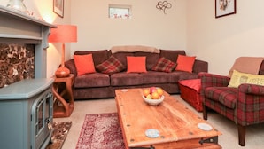 Ein gemütliches Wohnzimmer mit einem braunen Sofa, roten Kissen, einem karierten Sessel, einem Holztisch mit Obst und einer Vintage-Lampe.