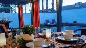 An outdoor dining area with a wooden table set for four, surrounded by chairs. There is a grill and lounge chairs in the background.