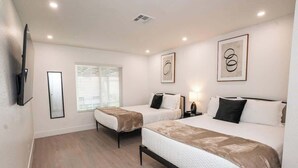 A hotel room with two beds, a TV, a window with blinds, and artwork on the walls.