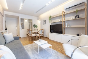 Una sala moderna con sofá, mesa de centro y televisor. Hay un comedor con sillas y una mesa. El piso es de madera y las paredes son blancas.