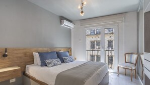 A bedroom with a wooden headboard, a bed with white bedding and blue pillows, a bedside table, a chair, and a balcony with a view of buildings. - Renovated 2BR in Recoleta | Balcony & Smart Lock (Recoleta)