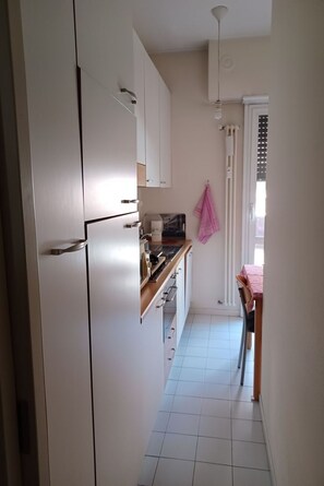 A narrow kitchen with white cabinets, a countertop, and a window with a curtain. - BnButler - Mussi, 10 (Milano)