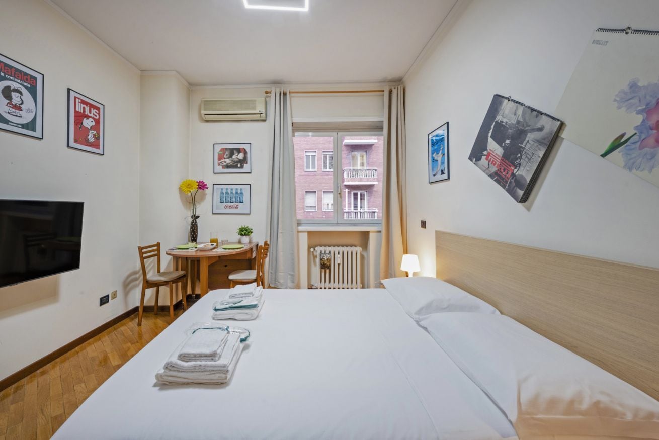 A hotel room with a bed, a TV, a desk with a chair, a window with a view of buildings, and artwork on the walls.
