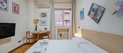 A hotel room with a bed, a TV, a desk with a chair, a window with a view of buildings, and artwork on the walls.