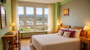 Habitación de hotel con una cama grande, un escritorio, una silla y vistas a un lago y montañas.