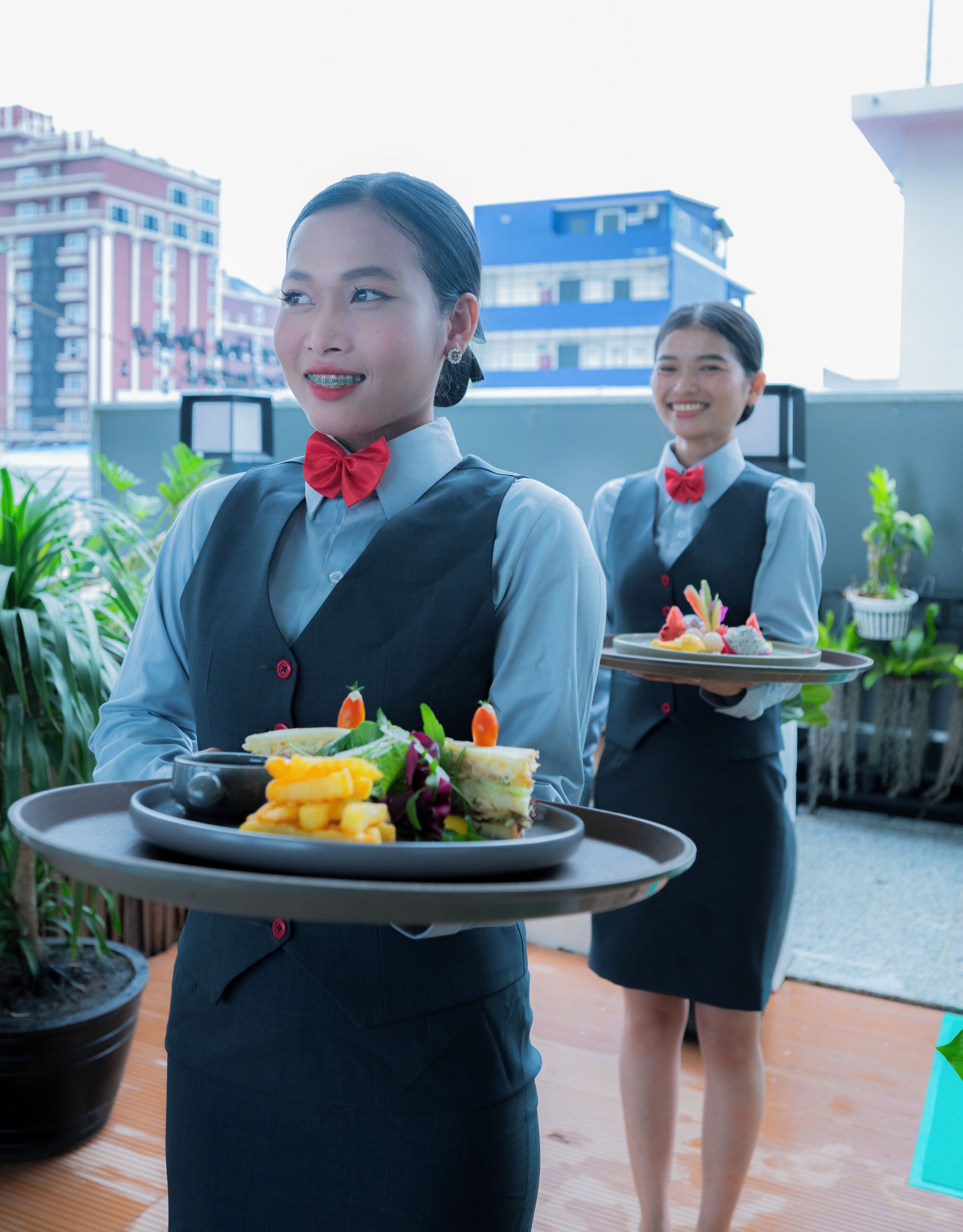 A person holding a tray with food, another person in the background, and a cityscape with buildings.