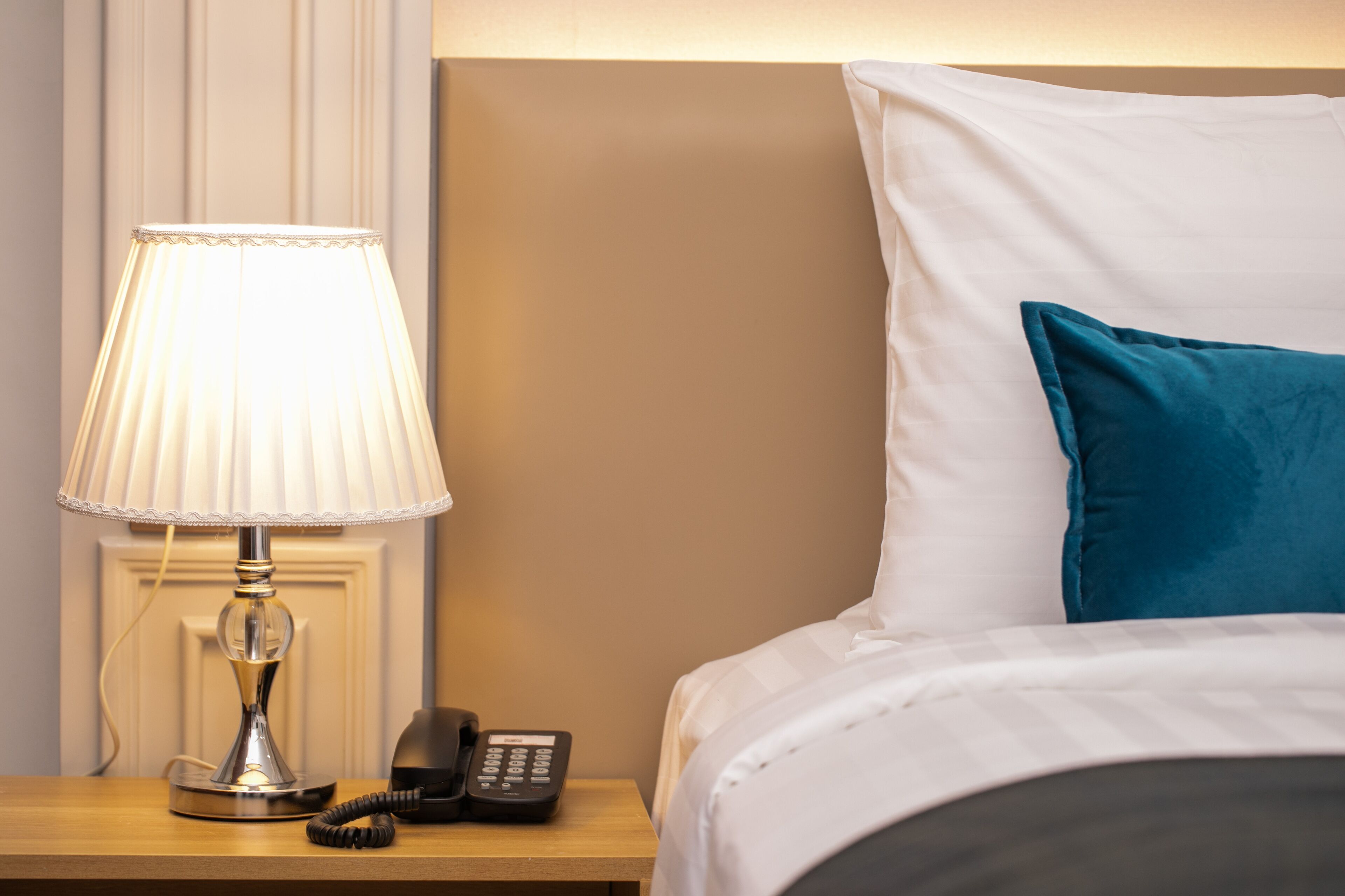 Una cama bien hecha con un almohadón blanco y azul turquesa, una lámpara de noche y un teléfono sobre una mesita de madera.