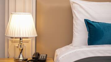 Una cama bien hecha con un almohadón blanco y azul turquesa, una lámpara de noche y un teléfono sobre una mesita de madera.
