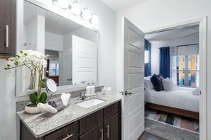 A modern bathroom with a large mirror, a sink, and a shower. There is a bed with a view of the city in the adjacent room. - Resort Style 1BR Near Lackland & Medical Center - Pool Gym Parking Pets OK (San Antonio)