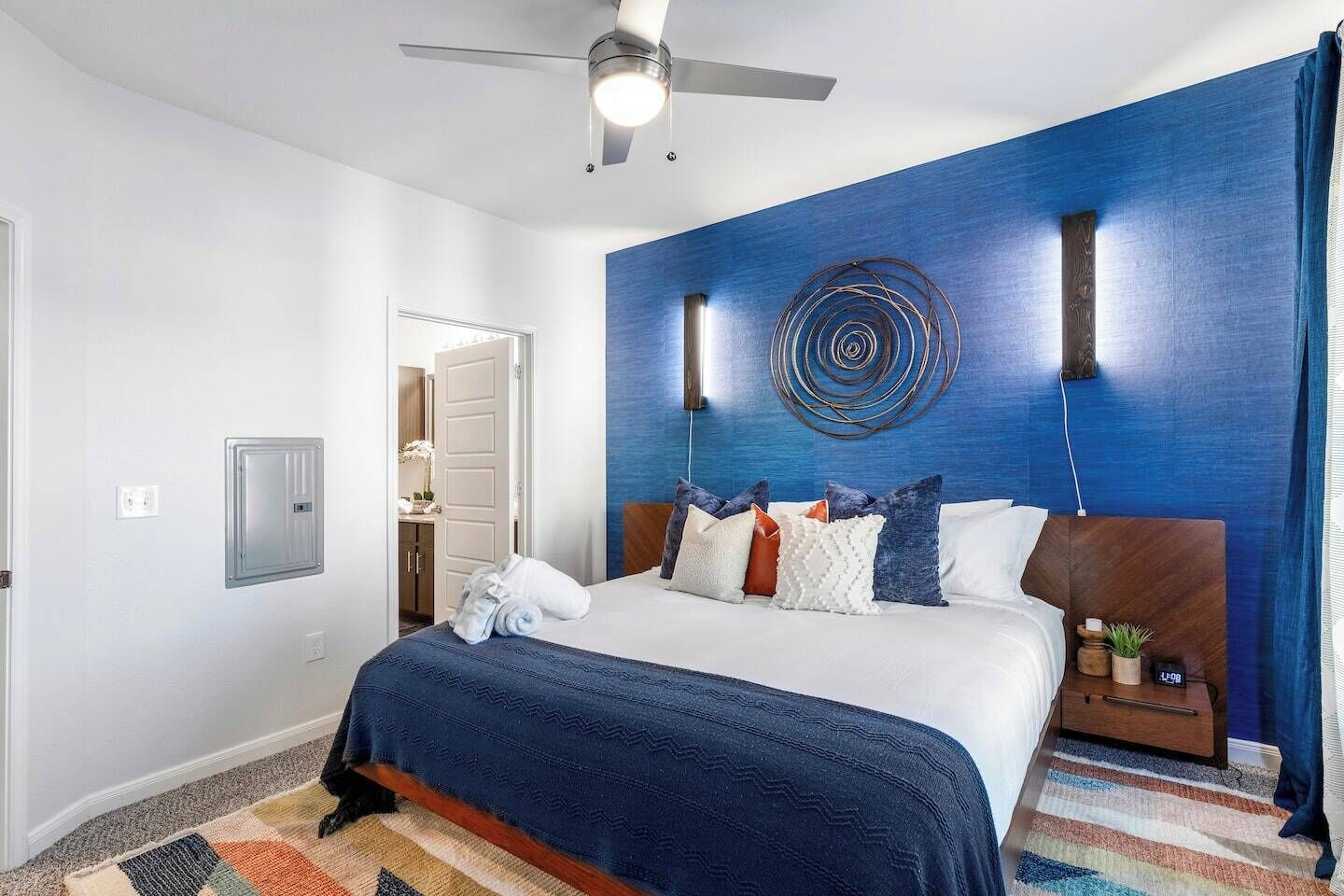 A hotel room with a bed, a ceiling fan, a wall-mounted light, a power outlet, and a door leading to a bathroom.