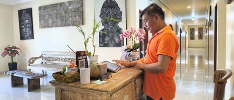 A reception desk with a wooden counter, a QR code, and a digital tablet. There are two potted plants, a white bench, and a wall with artwork.