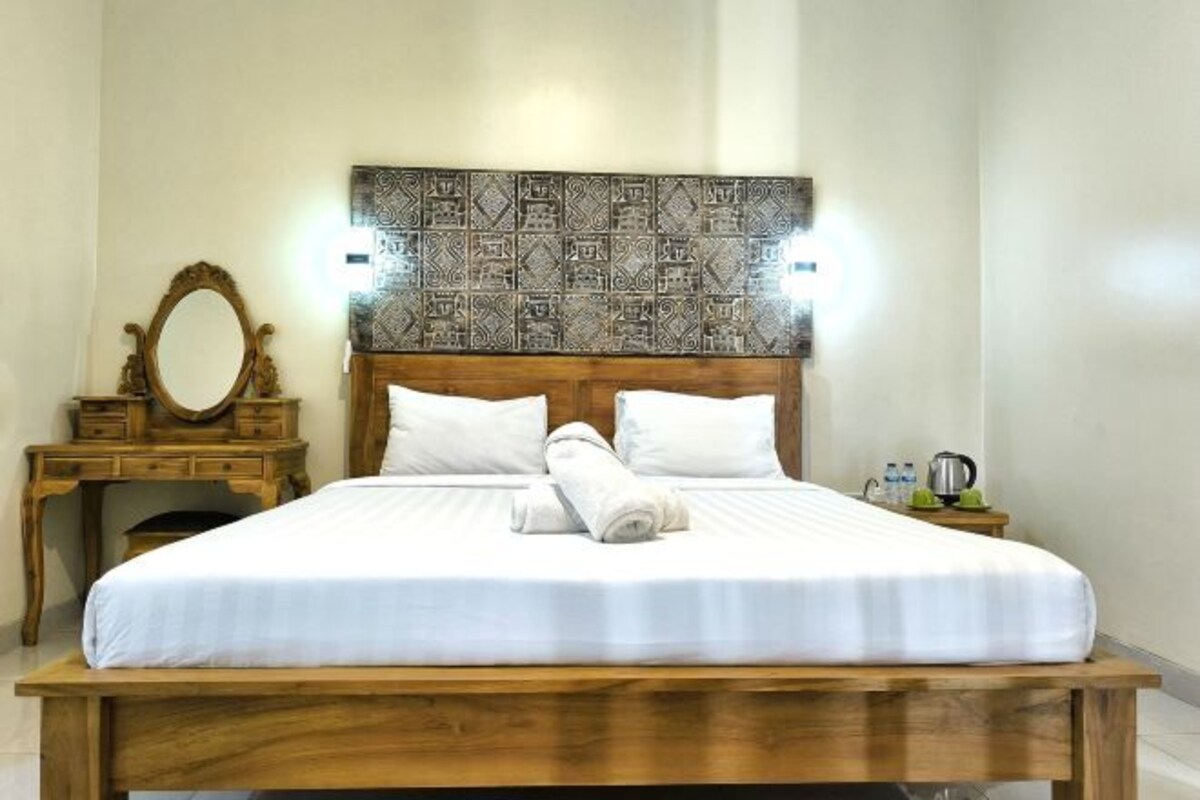 Una cama bien hecha con ropa blanca, un cabecero de madera con diseño estampado, una mesita de noche con un espejo y una tetera, y una pequeña mesa con una lámpara.