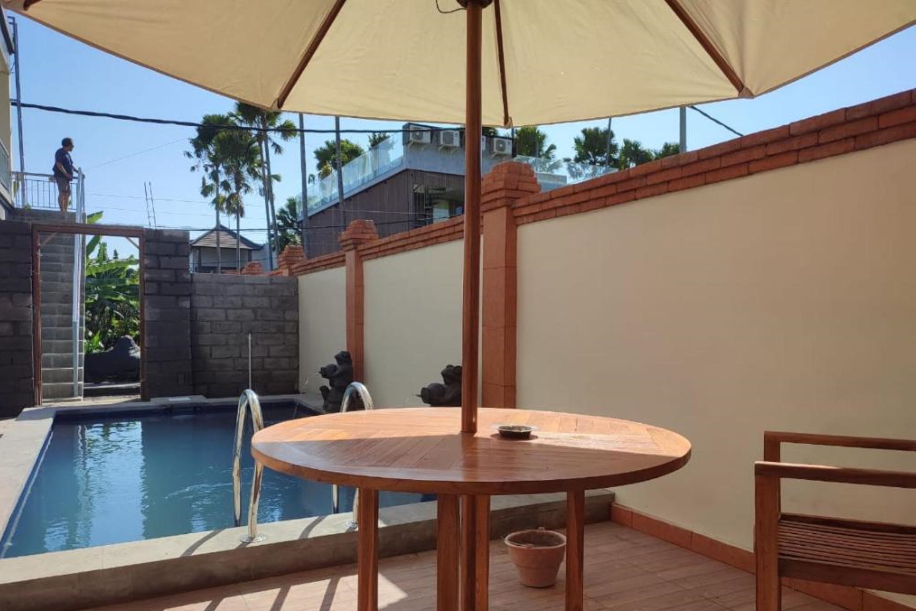 A pool area with a round wooden table and a chair, a ladder leading to a balcony, and a brick wall.