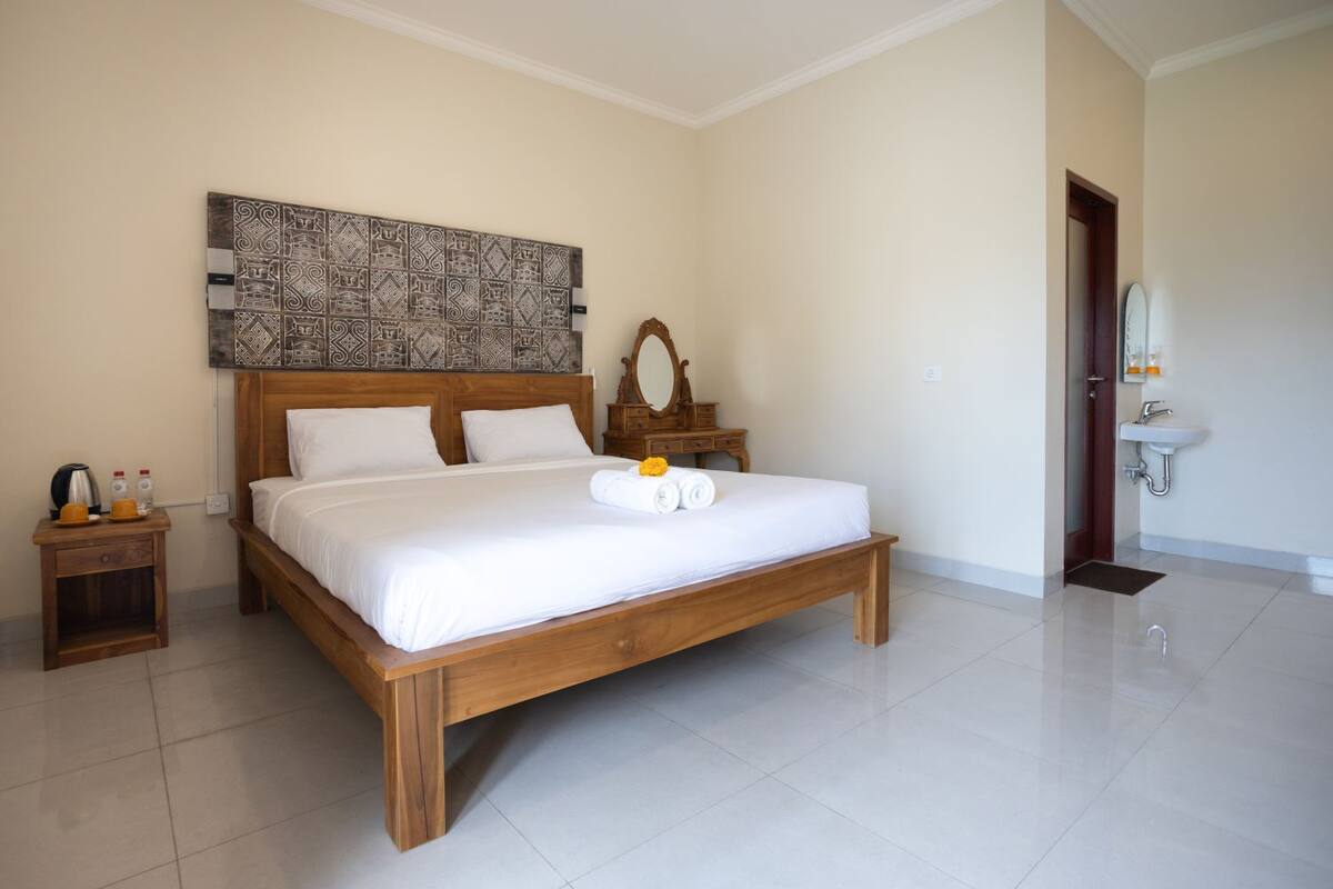 A hotel room with a wooden bed, a nightstand with a kettle and bottles, a mirror, and a bathroom with a sink and mirror.