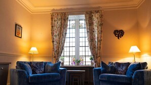 Una habitación con dos sillones azules, un tapete estampado, un radiador y una ventana con cortinas de flores.