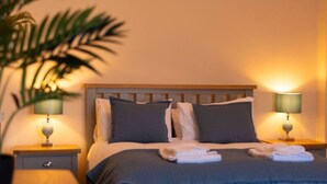 Una cama bien hecha con edredón gris y blanco, dos lámparas de noche, una mesita de noche de madera y una planta en maceta.
