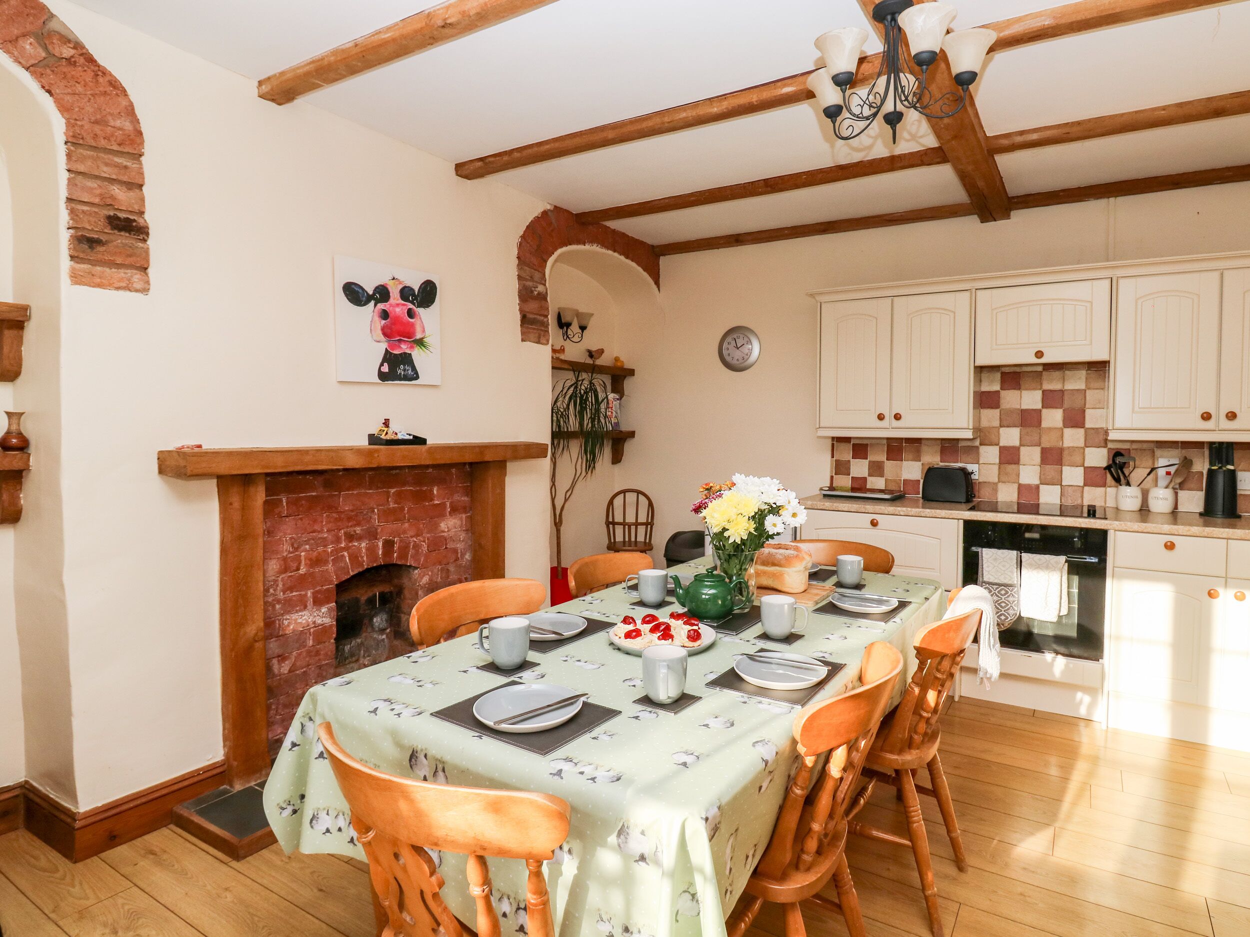 A dining area with a brick fireplace, wooden chairs, a table set for two, and a kitchen in the background.
