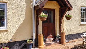 A house with a wooden front door, a small porch, and hanging flower baskets. - Twyford Farm Cottage (Tiverton)